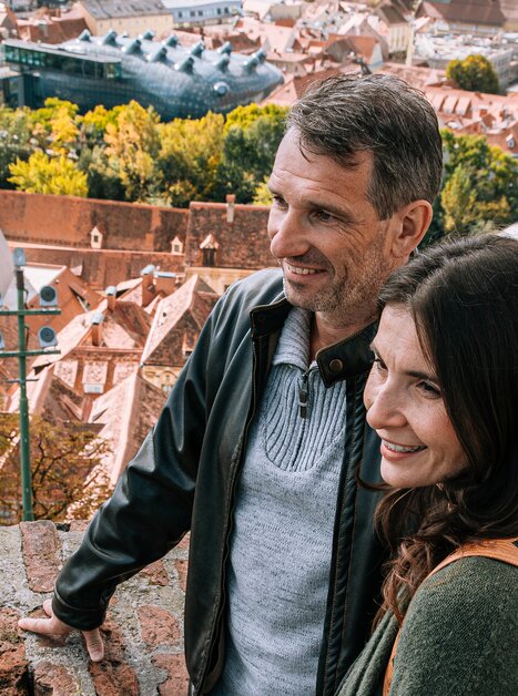 Paar genießt die Aussicht auf Graz. Im Hintergrund ist das Kunsthaus und die wunderschöne rote Dachlandschaft zu sehen. | © Graz Tourismus