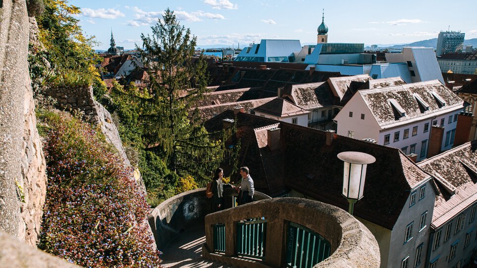 Blick auf Graz vom Schlossbergsteig mit den roten Dächern im Hintergrund. Ein Pärchen geht den Schlossbergsteig hinauf. | © Graz Tourismus
