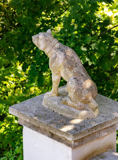 A stone sculpture of a dog on a pedestal, surrounded by green foliage. | © Graz Tourismus - Harry Schiffer