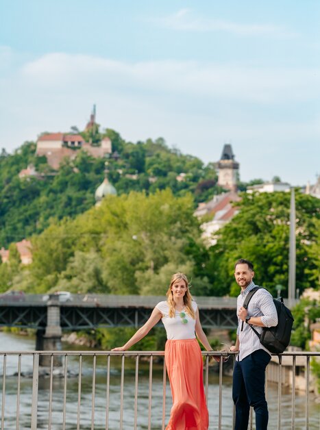 Coppia vicino alla Mur con Torre dell'Orologio di Graz sullo sfondo. | © Graz Tourismus - Mias Photoart