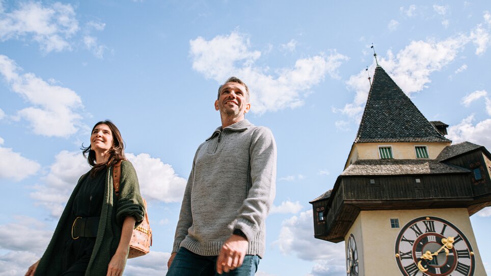 Couple walking in front of the Graz clock tower under a blue sky. | © Graz Tourismus