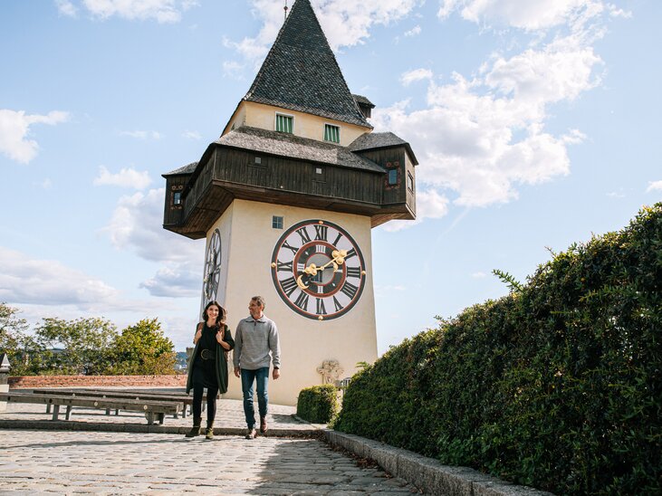 Couple walking in front of the Graz clock tower under a blue sky. | © Graz Tourismus