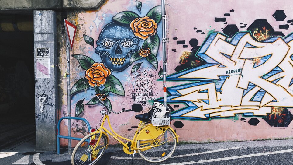 Gelbes Fahrrad vor einer Wand mit farbenfrohem Graffiti von einem Totenkopf und Rosen. | © Adeline Gressin