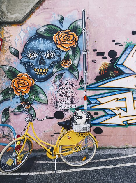 Gelbes Fahrrad vor einer Wand mit farbenfrohem Graffiti von einem Totenkopf und Rosen. | © Adeline Gressin