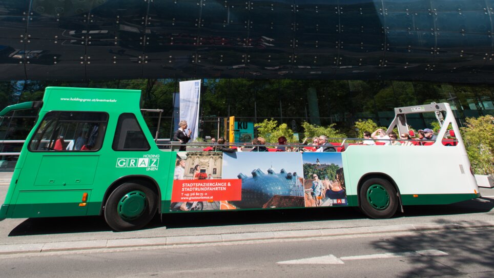 A cabrio bus passes by Kunsthaus Graz, blue sky and green trees in the background. | © Graz Tourismus - Harry Schiffer