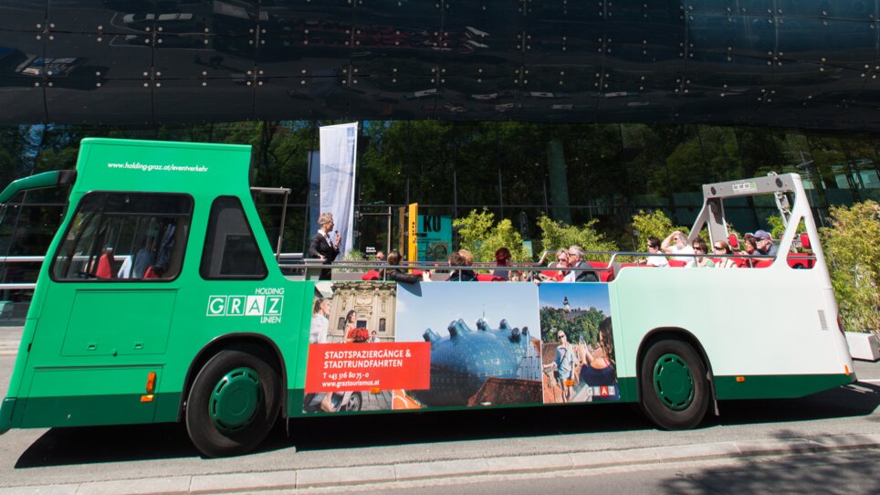 Ein Cabriobus fährt am Kunsthaus Graz vorbei, blauer Himmel und grüne Bäume im Hintergrund. | © Graz Tourismus - Harry Schiffer