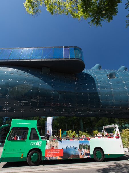 Ein Cabriobus fährt am Kunsthaus Graz vorbei, blauer Himmel und grüne Bäume im Hintergrund. | © Graz Tourismus - Harry Schiffer
