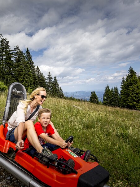 Eine Mutter und ihr Sohn fahren auf einer Sommerrodelbahn in den Bergen. | © Region Graz - Tom Lamm