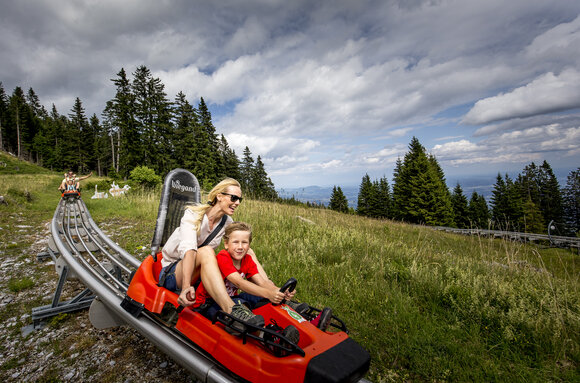 Eine Mutter und ihr Sohn fahren auf einer Sommerrodelbahn in den Bergen. | © Region Graz - Tom Lamm