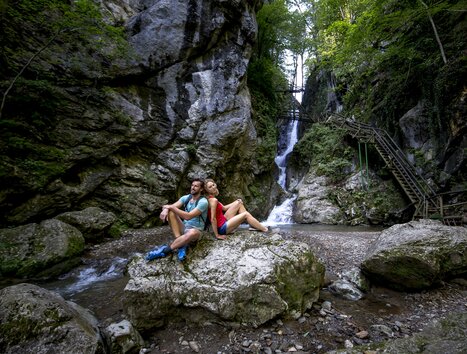 Ein Paar sitzt auf einem Felsen neben einem Wasserfall in der Kesselfallklamm. | © Region Graz - Tom Lamm
