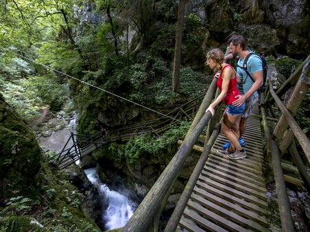 Ein Paar steht auf einer Holzbrücke und schaut auf den Fluss in der Kesselfallklamm. | © Region Graz - Tom Lamm