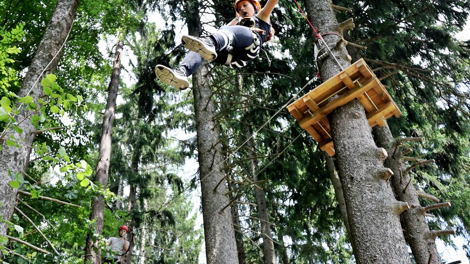 Junge schwingt an einem Seil zwischen Bäumen, während andere Kletterer in der Nähe sind. | © Kletterpark Schöckl - Oberlaender