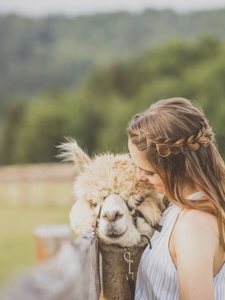 Eine Frau steht in der Natur und umarmt ein Alpaka freundlich. | © dieSonne