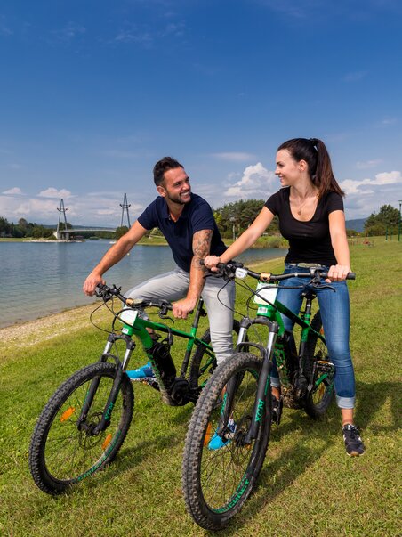 Paar auf Fahrrädern am Ufer des Schwarzlsee, strahlender Himmel. | © Region Graz - Harry Schiffer