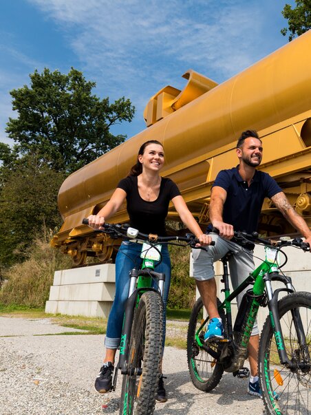 Zwei Personen fahren mit Fahrrädern vor einer goldenen Skulptur im Österreichischen Skulpturenpark. | © Region Graz - Harry Schiffer