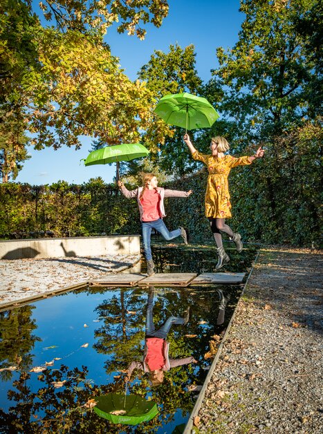 A girl and a woman jump over a water surface with green umbrellas. | © Region Graz - Mias Photoart