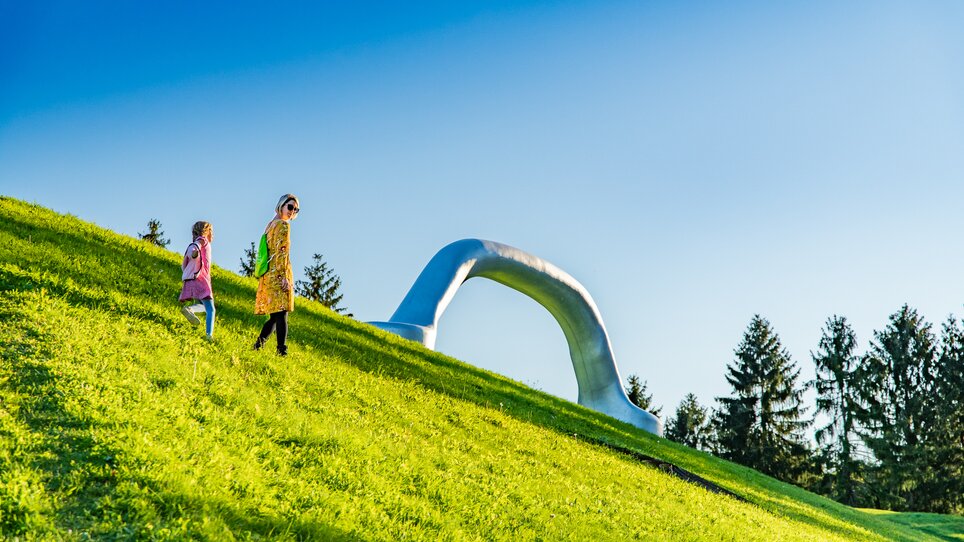 Eine Mutter und ihre Tochter spazieren durch den Österreichischen Skulpturenpark bei Graz, im Hintergrund ist der riesige Griff eines Koffers zu sehen. | © Region Graz - Mias Photoart