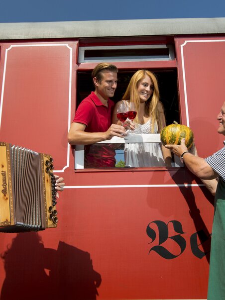 A couple on the Flascherlzug in Stainz receives wine and a pumpkin through the train window from a local farmer, while a woman in traditional dress plays a Styrian accordion on the platform. | © Steiermark Tourismus - Harry Schiffer