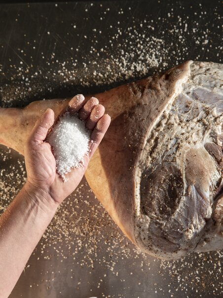 Close-up of hands sprinkling salt over a raw ham as part of the traditional curing process | © Vulcano