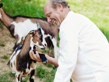 A smiling man is happily interacting with a goat. | © Zotter Schokolade