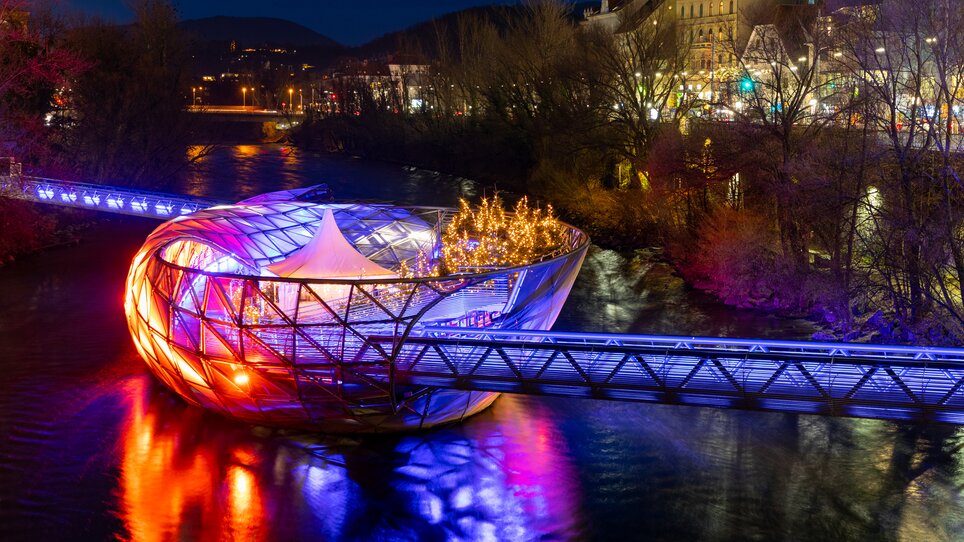 Die Murinsel in Graz erstrahlt in bunten Lichtern über dem Fluss Mur. | © Graz Tourismus - Harry Schiffer I Lichtkonzept: Ochoresotto  