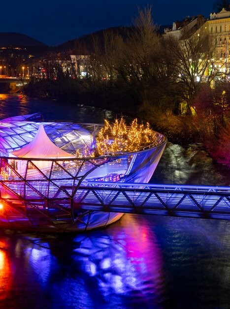 Die Murinsel in Graz erstrahlt in bunten Lichtern über dem Fluss Mur. | © Graz Tourismus - Harry Schiffer I Lichtkonzept: Ochoresotto  