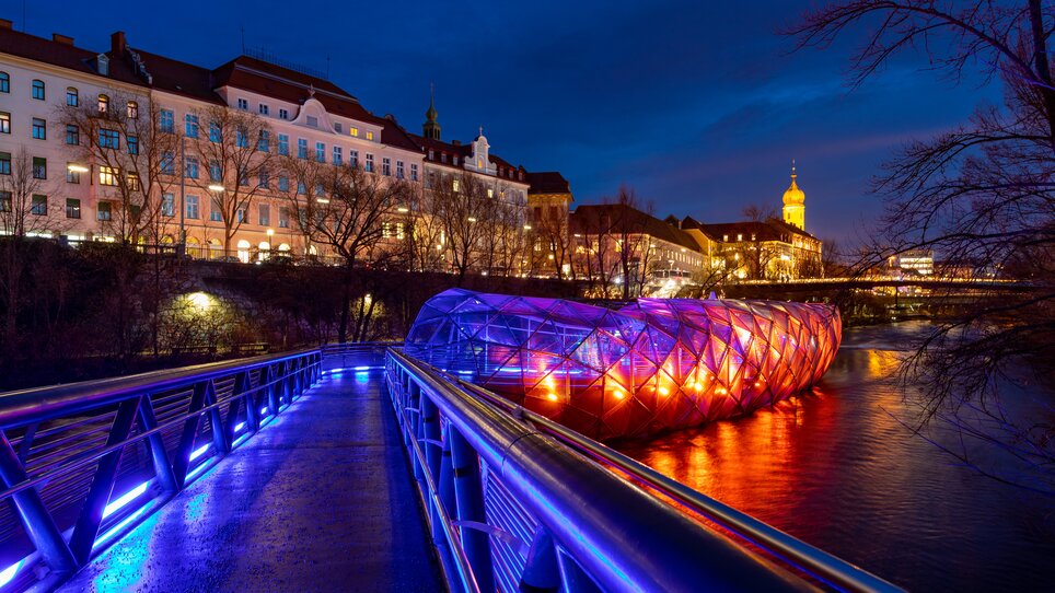 Beleuchtete Murinsel in Graz bei Nacht mit Blick auf die Stadt. | © Graz Tourismus - Harry Schiffer I Lichtkonzept: Ochoresotto  