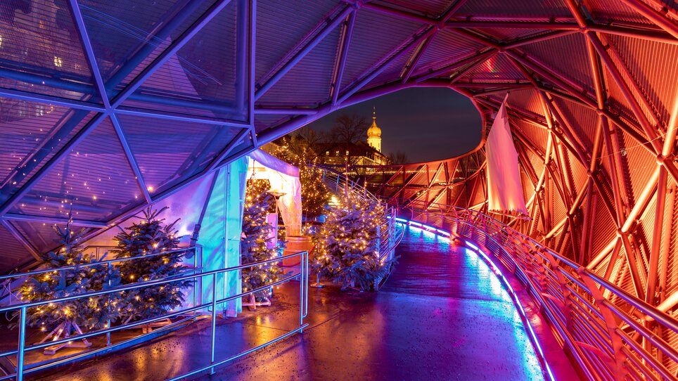 Futuristischer Weg mit bunten Lichtern und Weihnachtsbäumen auf der Murinsel in Graz. | © Graz Tourismus - Harry Schiffer I Lichtkonzept: Ochoresotto  