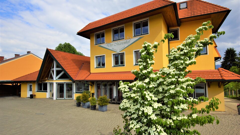 The image shows a charming yellow building with a red roof and blooming plants. | © Der Marienhof