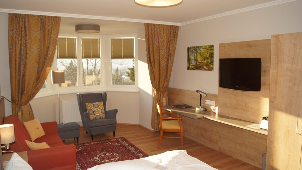 The image features a bright, comfortable room with windows, a seating area, and modern furnishings. | © Der Marienhof