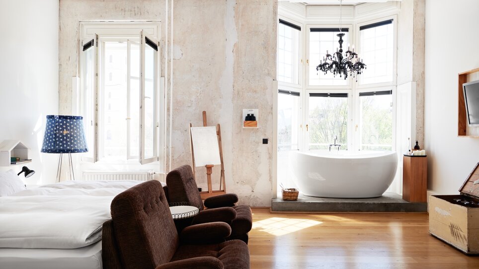 Helles Schlafzimmer mit großem Fenster, einer freistehenden Badewanne, zwei Stühlen und einem Fernseher. | © Weitzer Hotels