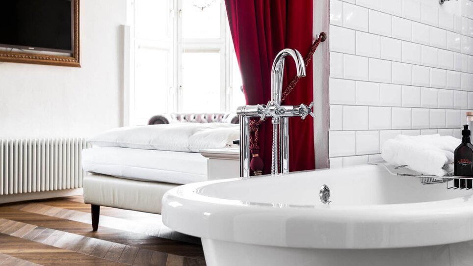 Ein Badezimmer mit freistehender Badewanne und rotem Vorhang, Schlafzimmer im Hintergrund.