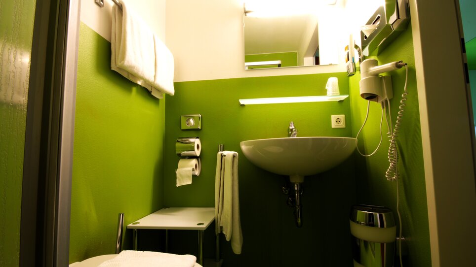 Bagno compatto con pareti verdi, lavabo e asciugamani. | © Hotel Greenrooms