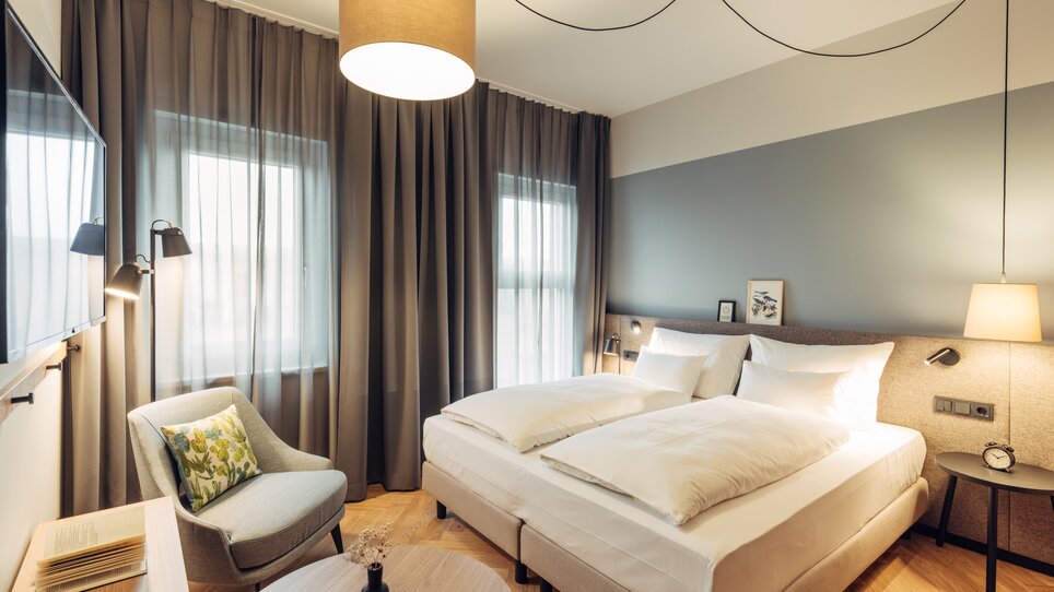 Helles Schlafzimmer mit zwei Betten, Fernseher und Stuhl im harry's home Graz-Smart City. | © harry’s home hotels - Daniel Zangerl