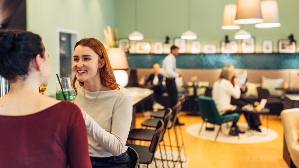 Zwei Frauen genießen Getränke in der modernen Bar des harry's home Graz-Smart City, während andere Gäste sich entspannen. | © harry’s home hotels - Daniel Zangerl