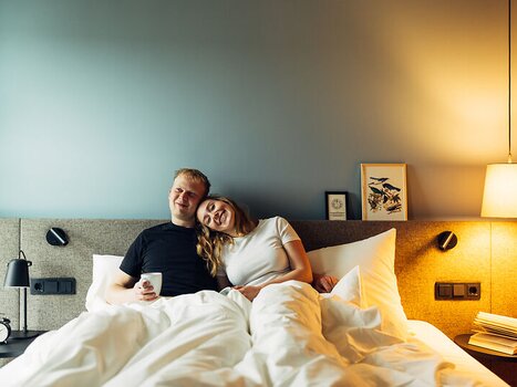 A couple sits in bed, happy and relaxed, surrounded by white blankets. | © Daniel Zangerl