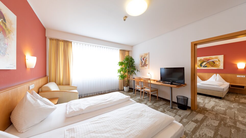 Bright double room with two beds, TV and plants and a passageway to a smaller room. | © Harry Schiffer