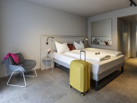 Camera d'albergo luminosa con letto matrimoniale, valigia gialla e sedia. | © Hotel Ibis Styles Graz Messe