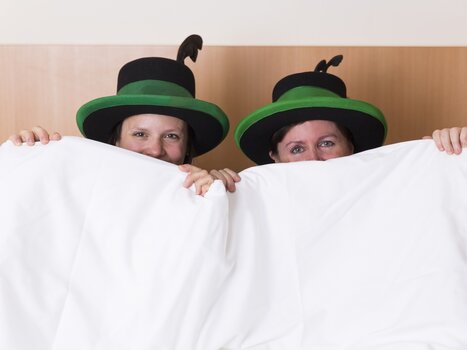 Due persone con cappelli verdi siedono nel letto tenendo su una coperta. | © Hotel Kern Buam