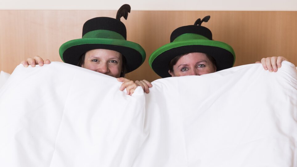 Zwei Personen mit grünen Hüten sitzen im Bett und halten eine Decke hoch. | © Hotel Kern Buam