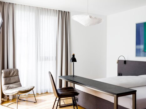 Bright room with a large bed, table, chair, and stylish lamp. | © Lendhotel