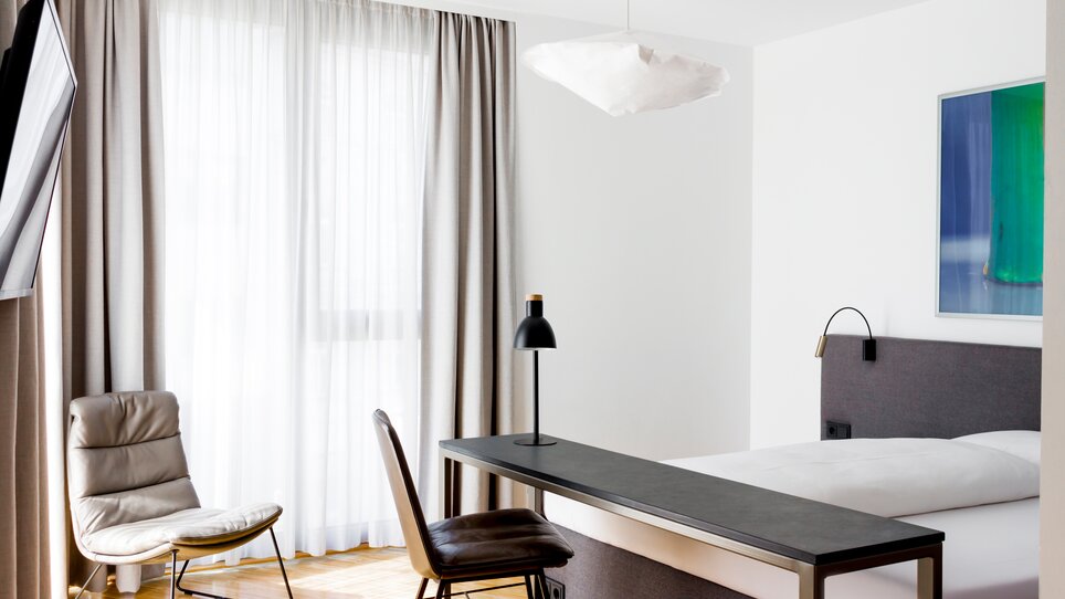 Stanza luminosa con letto grande, tavolo, sedia e lampada elegante. | © Lendhotel