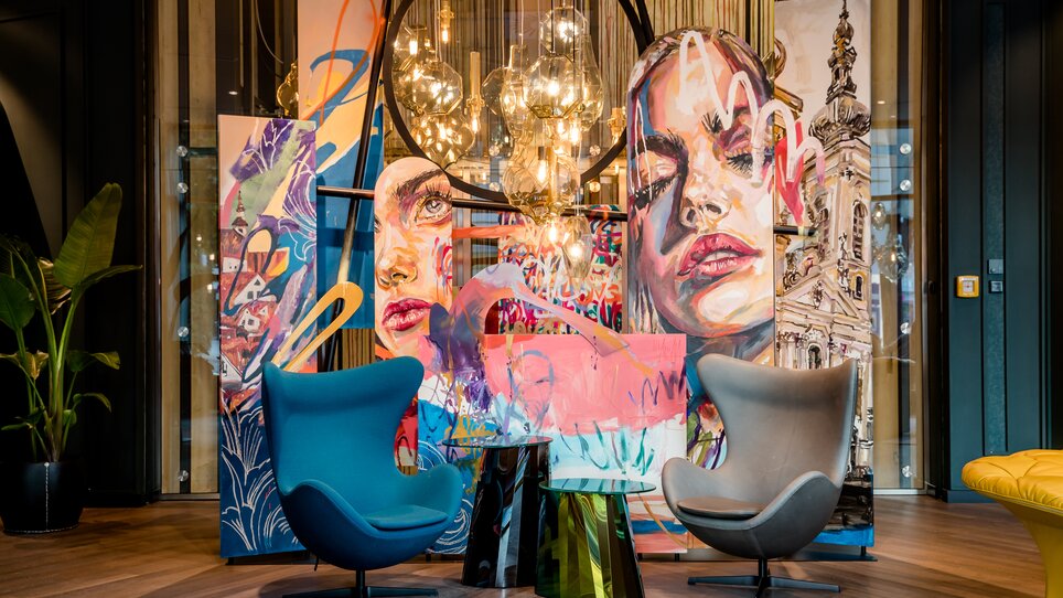 Helle Lounge mit blauen und grauen Sesseln, künstlerischer Wandgestaltung und leuchtenden Lampen. | © Motel One Austria GmbH