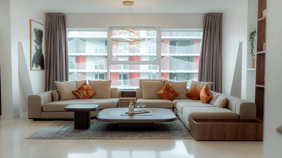 Bright living room with beige sofa, cushions, and large window. | © NED Hotel