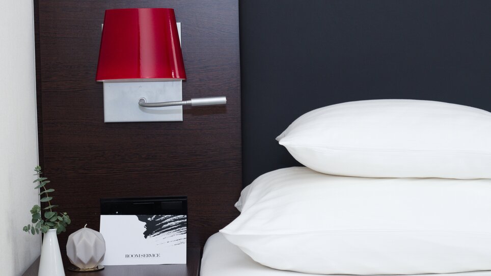 A nightstand with a red lamp and pillows | © PLAZA INN Graz City