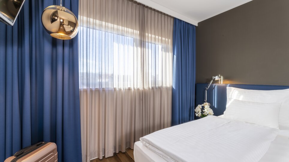 Bright double room with large windows and blue curtains. | © Plaza INN Graz