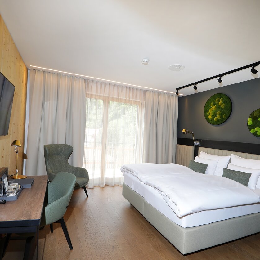 Modernes Doppelzimmer in Grautönen mit Bett, Schreibtisch, Fernseher und großer Fensterfront. An der Wand hängen mehrere grüne Deko-Elemente mit Moos. | © Hotel Steiermarkhof