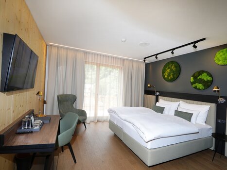Modernes Doppelzimmer in Grautönen mit Bett, Schreibtisch, Fernseher und großer Fensterfront. An der Wand hängen mehrere grüne Deko-Elemente mit Moos. | © Hotel Steiermarkhof
