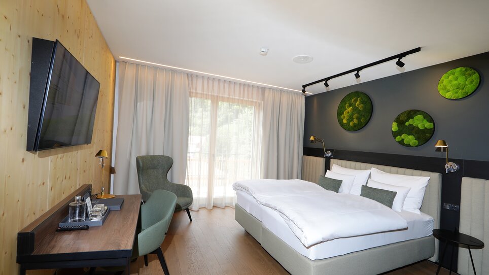 Modernes Doppelzimmer in Grautönen mit Bett, Schreibtisch, Fernseher und großer Fensterfront. An der Wand hängen mehrere grüne Deko-Elemente mit Moos. | © Hotel Steiermarkhof