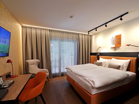 Modernes Doppelzimmer mit Bett, Schreibtisch, Fernseher und großer Fensterfront. Der Raum ist in warmen Beige- und Orangetönen gestaltet. | © Hotel Steiermarkhof